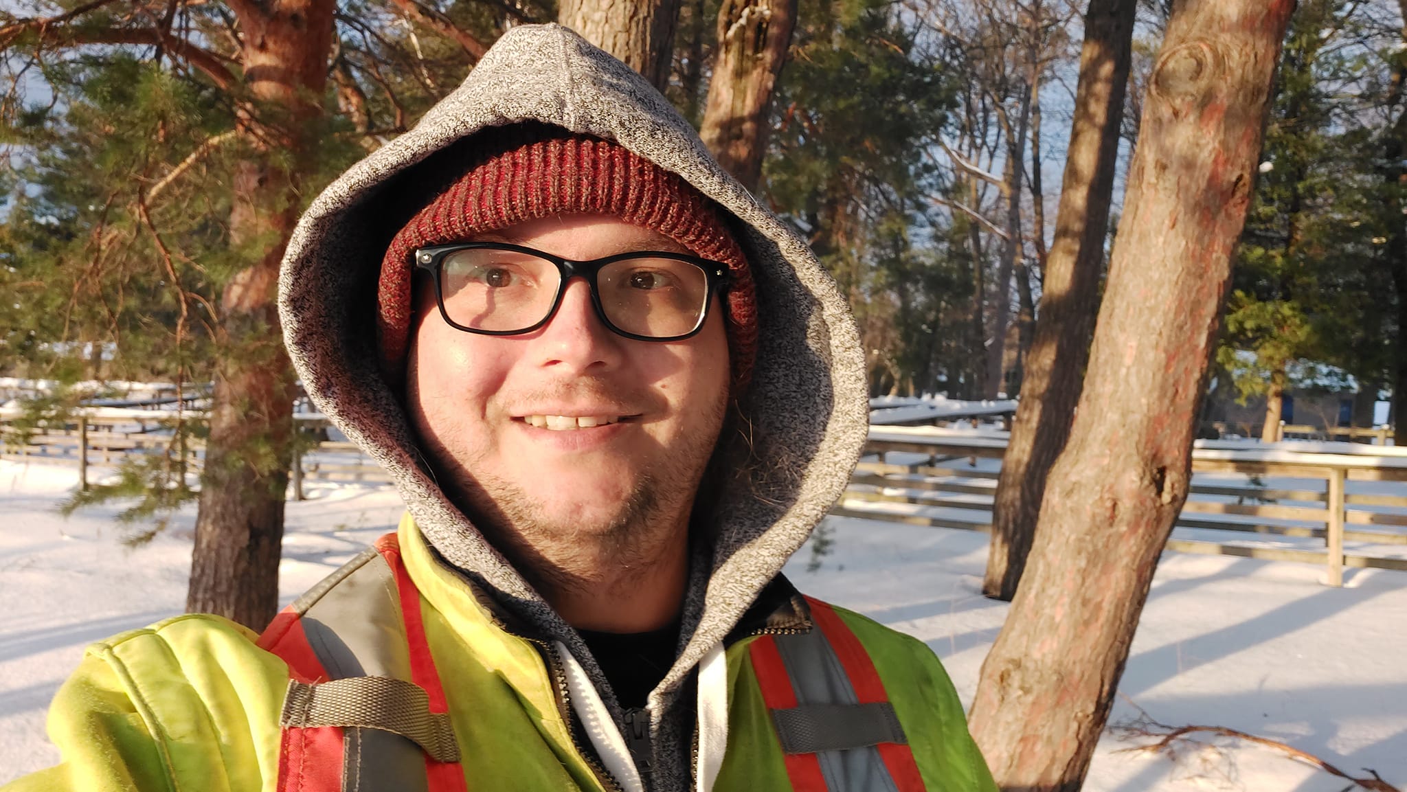 A man with green hair is wearing a high visibility winter coat, a red toque with a grey hood also visible, a boardwalk covered in snow is visible behind him